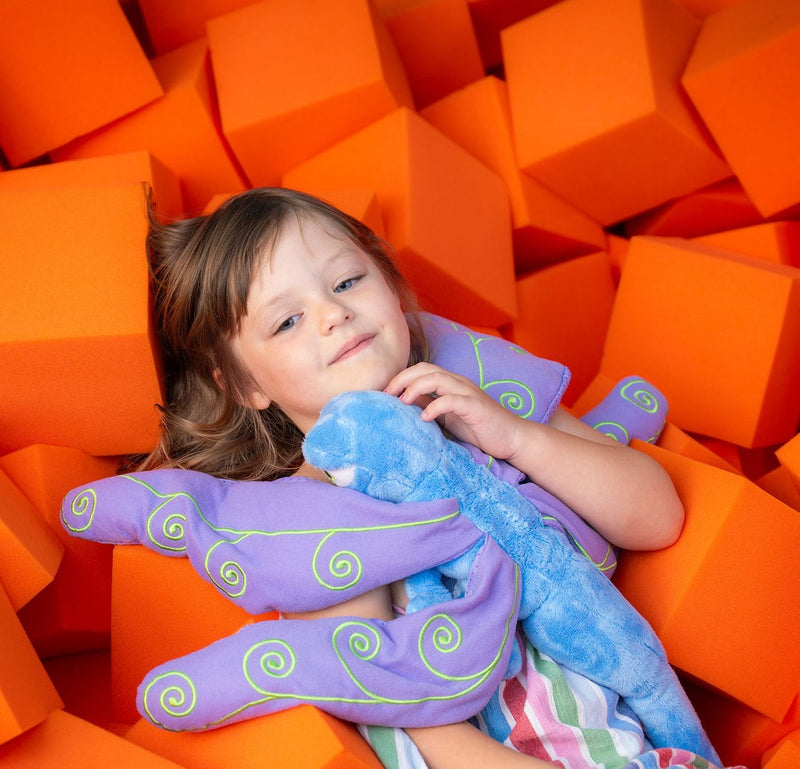 A child lying back in a foam block pit, holding Flutter against her chest.