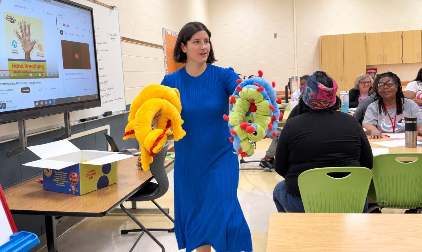 Sarah Habib holding two Calmee plush caterpillars while leading a teacher training session in a classroom.