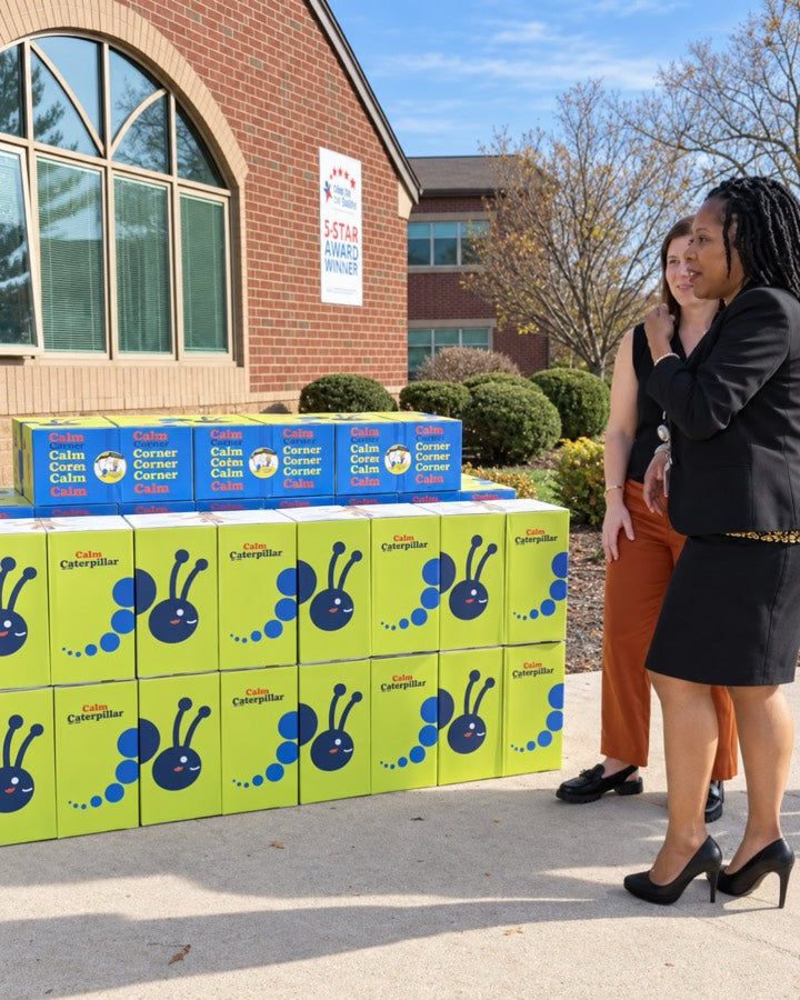 Two school staff members standing beside stacked Calm Caterpillar and Calm Corner kit boxes outside a school entrance.
