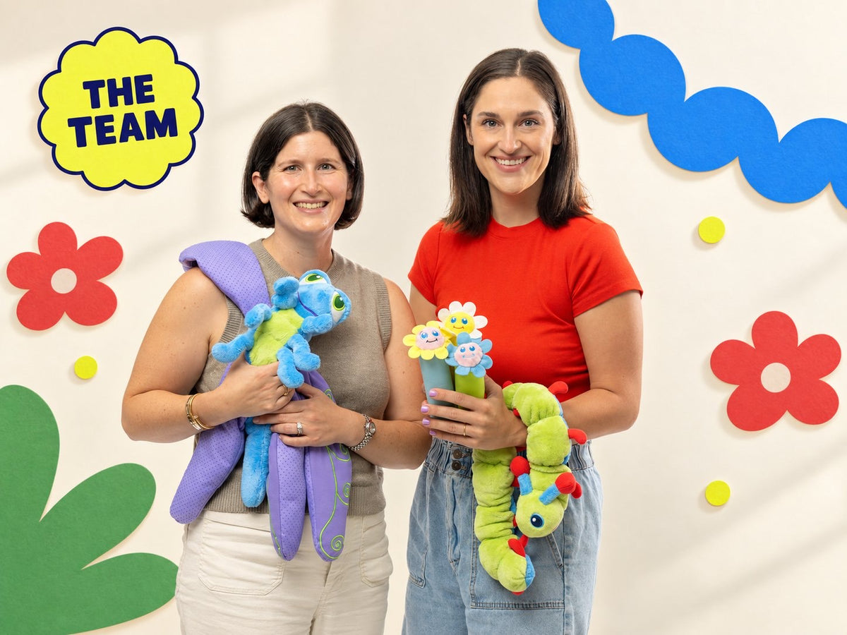 Two members of the Calm Caterpillar team holding Calmee, Flutter, and Bloomie in front of a paper-flower backdrop.
