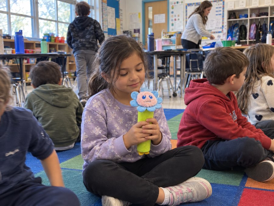 A girl sitting cross-legged on the classroom rug after recess, holding Bloomie the flower breathing tool in both hands while classmates settle around her.