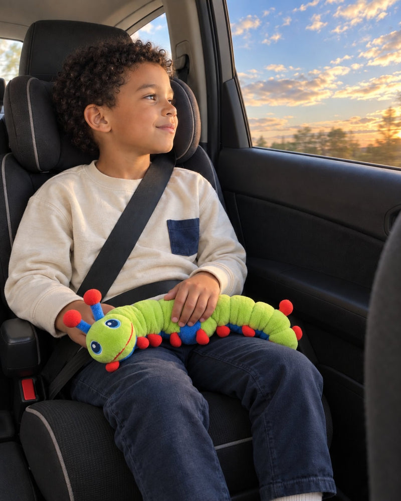 A boy buckled into a car seat at sunset, resting both hands on Calmee the Caterpillar across his lap