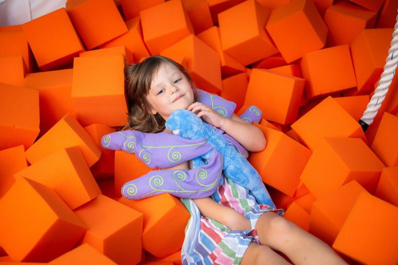 A girl lying back in a foam-block pit hugging Flutter against her chest, wings wrapped around her arms