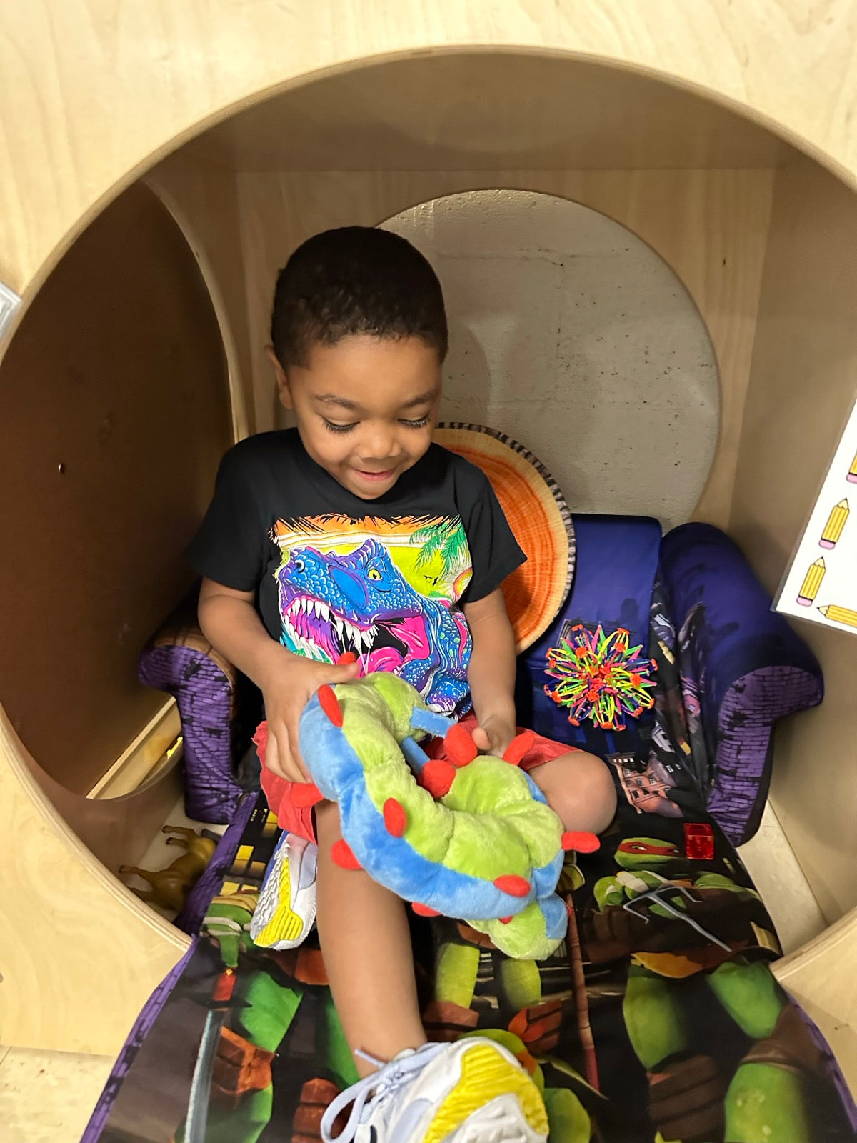 A boy sitting inside a wooden classroom calm corner pod, smiling and holding Calmee the Caterpillar across his knees