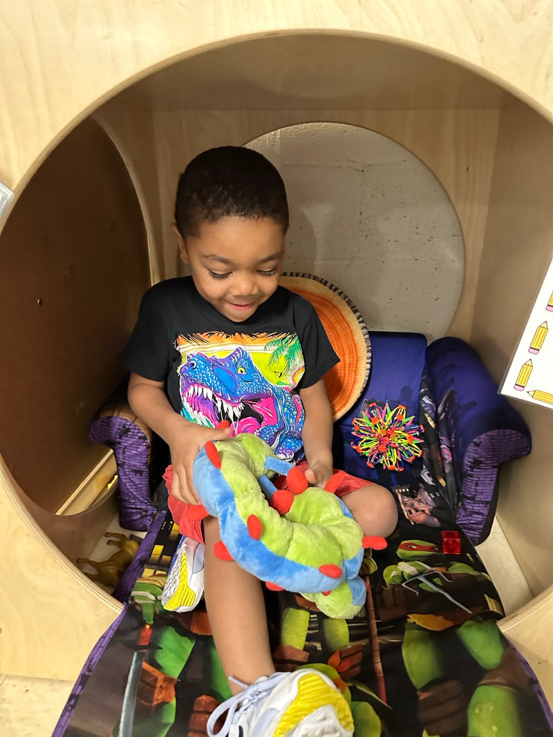 A boy sitting inside a wooden classroom calm corner pod, smiling and holding Calmee the Caterpillar across his knees