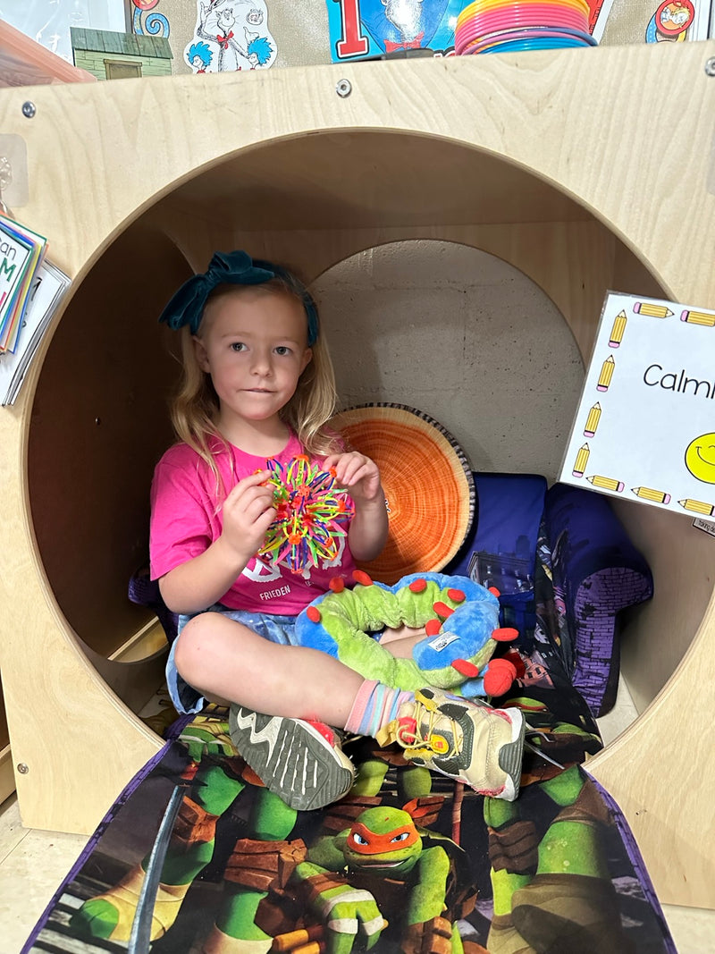 A girl sitting in the same calm corner pod, holding the expandable breathing ball open with Calmee in her lap