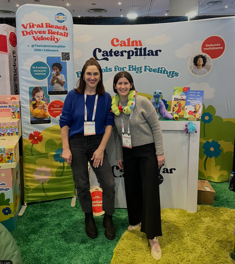 Kathryn and Sarah standing in the Calm Caterpillar Toy Fair booth, one wearing Calmee around her neck, in front of the Big Tools for Big Feelings backdrop.