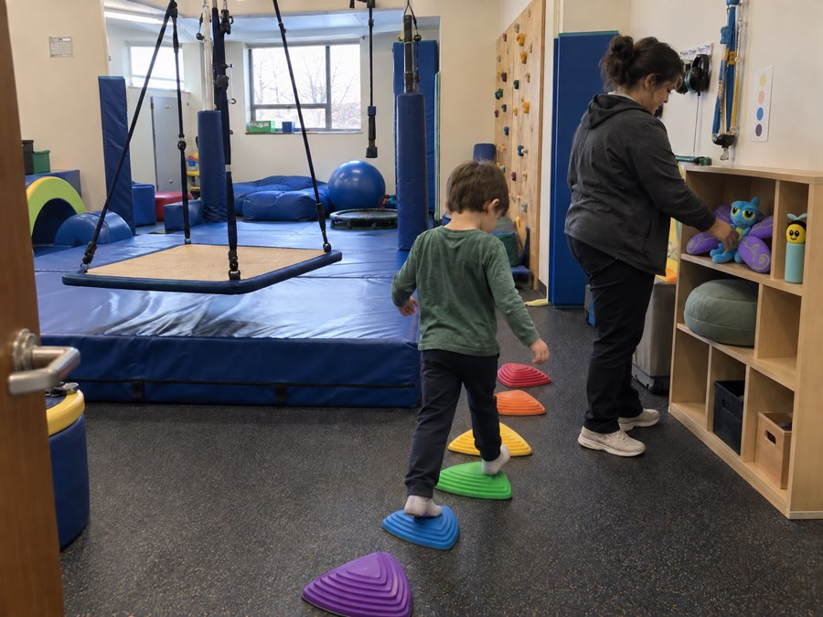 A child steps across stepping stones in a sensory gym with a platform swing, crash mat, and climbing wall behind them; a staff member sets Flutter and Buzzee on an open shelf beside floor cushions.