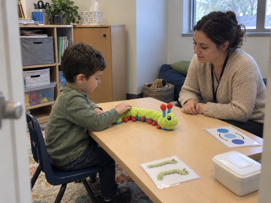 A child at a table in a session room rests a hand on a Calmee the Caterpillar plush while a practitioner sits beside them; visual cards and a supply bin are on the table.