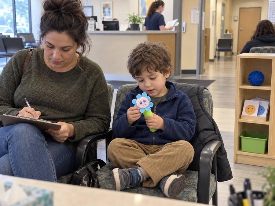 A child sits cross-legged in a clinic waiting area holding a Bloomie flower while a parent fills in a form beside them; a shelf nearby holds a spiky ball, feeling cards, and a storage bin.