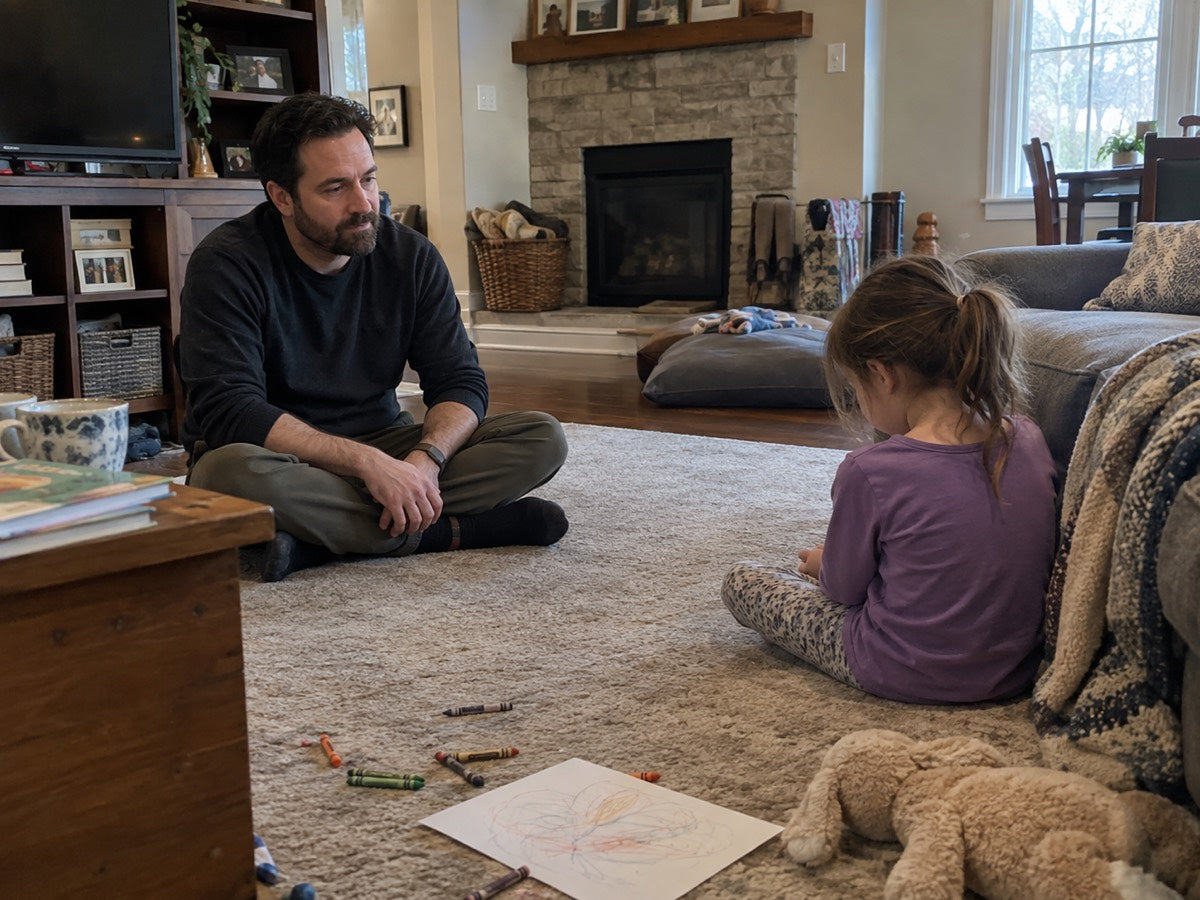 A parent sitting cross-legged on the living room floor a few feet from their child, calm and nearby.