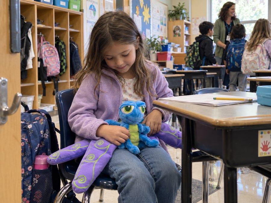 A girl sitting at her classroom desk at arrival, coat still on, holding Flutter the weighted dragonfly in her lap while classmates and a teacher come in behind her.