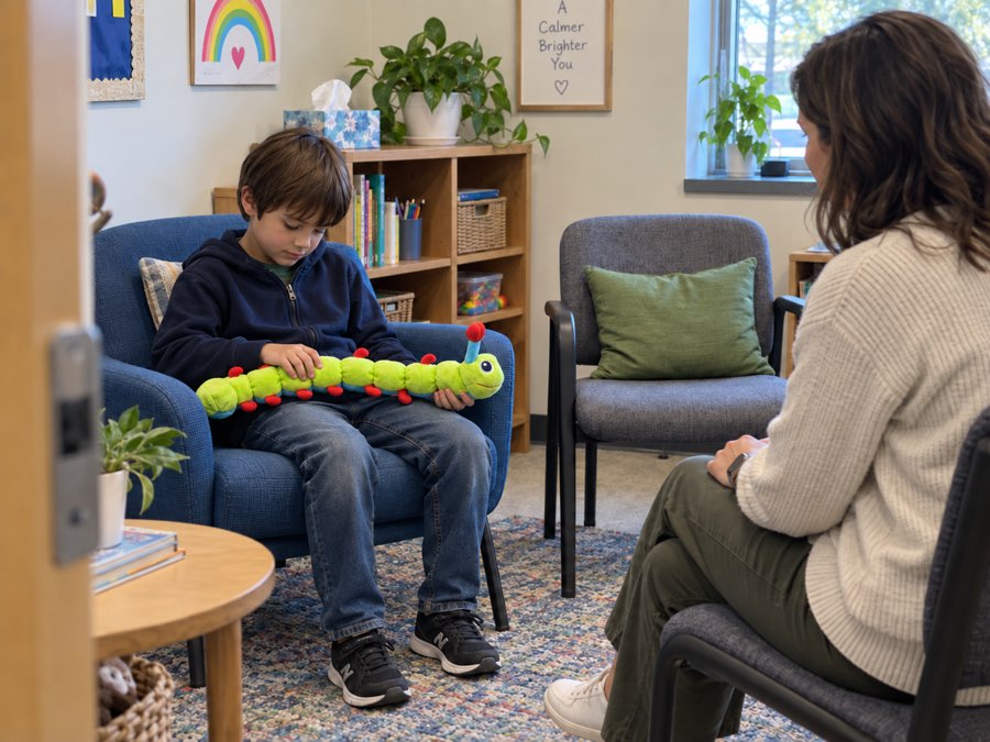 A boy sitting in an armchair in a counselor's office holding Calmee the caterpillar plush across his lap, with a counselor seated nearby.