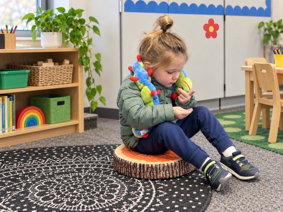 A young girl sitting on a tree-stump cushion on a classroom rug with Calmee the caterpillar around her neck.