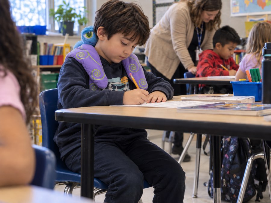 A boy working at his desk with Flutter the weighted dragonfly draped over his shoulders, pencil in hand, while a teacher helps another student behind him.