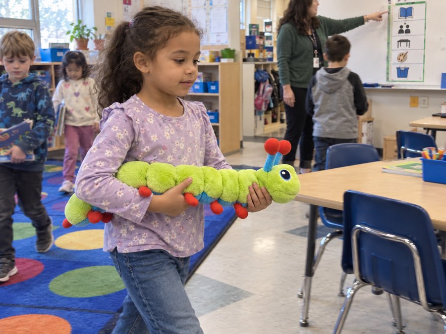 A girl carrying Calmee the caterpillar plush across the classroom rug during a transition, while a teacher points at a visual schedule and other children move to the next activity.