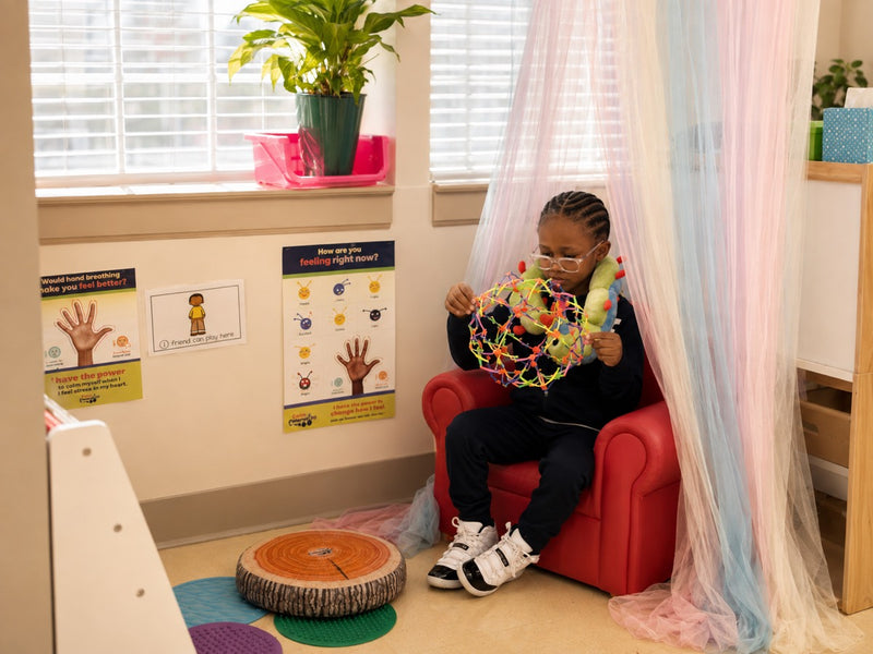 A child sitting in a classroom calm corner under a canopy, wearing Calmee around her neck and opening a sphere toy, with feelings posters on the wall